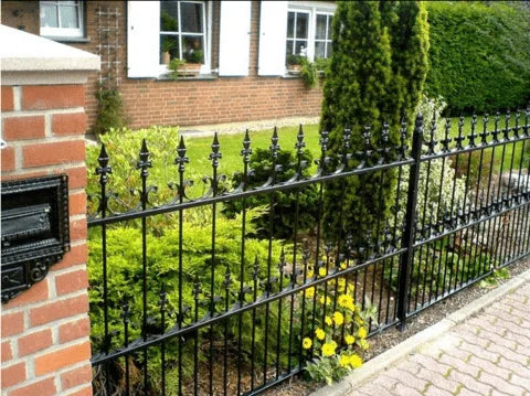 Valla de hierro forjado negro en un jardín con un pilar de ladrillo y una casa al fondo.